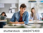 Small photo of Happy student taking notes while studying in high school. Satisfied young man looking at camera while sitting at desk in classroom. Portrait of college guy writing while completing assignment.