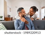Smiling ethnic woman hugging her husband on the couch from behind in the living room. Middle eastern man having fun with his beautiful young wife on the couch. Mid adult indian man with latin woman.