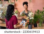 Small photo of Young african florist holding bouquet of fresh flower while customer making digital payment with smartphone while scanning QR code. Black saleswoman giving a bunch of flower to customer at botany shop