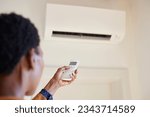 Black woman hand adjusting AC temperature with remote control to wall type air conditioning home cooling system. Hand of african woman holding remote controller directed towards the air conditioner.