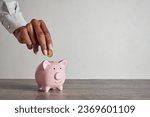 Small photo of Close up of black woman hand adding coin to a little pink piggy bank. African hand of businesswoman putting money into piggy bank for saving. Home finance and clever investment strategy concept.