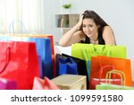 Worried shopaholic woman after multiple purchases in colorful shopping bags at home