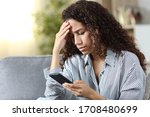 Worried latin woman reading on smart phone bad news sitting on a couch at home