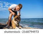 Small photo of Cheerful kind teenage guy with blond hair and comfortable leash in his hands plays and walks with his big fluffy multi-colored dog of Akina Inu breed, on sandy wild sea beach along Black Sea