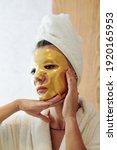 Small photo of Unsmiling woman in bathrobe with golden gel sheet mask on her face looking at herself in mirror, skincare and anti-aging treatment concept