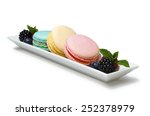 Small photo of Filled macaroons each of different color (blue, yellow, pink) prearranged on a rectangle plate decorated with blueberries, raspberries and mint leaves isolated on a white background.