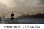 Black silhouette of a traditional barquentine or schooner barque tall ship cruising westwards the waters of Port Jackson in a very hazy afternoon-Harbour Bridge in the background. Sydney-NSW-Australia