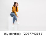 Small photo of Portrait of young Asian woman sitting on white box and keeps hand near mouth and whispers secret isolated over white background, Spreads rumors concept