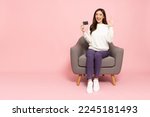 Small photo of Young elegant Asian woman showing credit card, ok sign and sitting on armchair isolated on pink background