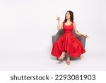 Happy young Asian woman in red dress sitting on armchair and pointing to empty copy space isolated on white background