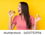 Young brunette woman over isolated yellow background holding colorful French macarons and eating it