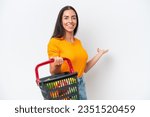 Small photo of Young caucasian woman holding a shopping basket full of food isolated on white background extending hands to the side for inviting to come