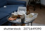 Small photo of Close up of messy table, leftover food, spilled drinks, dirty dishes in empty living room, bottle of beer and napkins on blue sofa. Unorganized apartment of person with sever depresion having rubbish