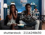 Small photo of Shocked african american couple watching tragic news on television program while sitting at home. Terrified people reacting bad while watching horror movies sitting alone on couch.