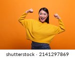 Small photo of Positive optimistic woman raising arms while feeling strong and confident on orange background. Motivated triumphant girl celebrating victory and feeling optimistic and independent.