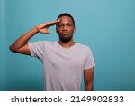 Small photo of Young man doing military salute with hand over forehead, advertising honor and trustworthy respect in front of camera. Proud adult showing army soldier hello gesture in studio. Veteran greeting