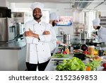 Small photo of Gastronomy expert standing in restaurant professional kitchen with arms crossed while smiling at camera. Confident head chef wearing cooking uniform while preparing ingredients for dinner service.