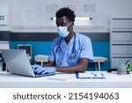 Small photo of Nurse wearing facemask while using laptop to analyze patient documents while sitting at desk in general practitioner cabinet. Clinic nurse verifying medicine expiry date, ingredients and dosage