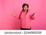 Small photo of Portrait of doubtful african american woman posing on pink background in studio while making indecisive facial expression. Young adult shrugging shoulders doing i dont know gesture