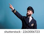 Small photo of Serious captain pointing at sky with index finger, wearing professional aviation uniform, plane pilot looking at camera. Confident aviation academy aviator standing, studio medium shot