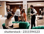 Small photo of Hotel concierge assisting guests in lobby, helping with check in process at reception front desk. Bellboy carrying trolley bags and luggage to room, giving luxury service at modern resort.