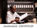 Small photo of African american prosecutor reading file, preparing for case in police office with archival records. Law enforcement professional analyzing forensic expertise folder at night time