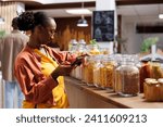 Small photo of African American woman grasping her smartphone with focused attention on the glass jars containing a variety of pasta. Using mobile device, young lady offers modern and efficient shopping experience.