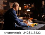 Small photo of Professional elderly tailor working with sewing materials to customize bespoken sartorial garments for comissioning client in atelier shop. Senior couturier manufacturing clothing in workspace