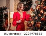 Happy beautiful young woman in holiday red dress decorating Christmas Tree with sparking balls and holds glass of champagne preparing for holiday. New Year concept. Merry Christmas 