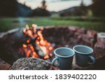 Campsite With Fire Pit and Two Tin Cups with hot tea. Burning Campfire with mountain landscape with evening sunset sky over the forest and hills.