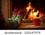 Feet in woollen socks by the Christmas fireplace. Woman relaxes by warm fire with a cup of hot drink and warming up her feet in woollen socks. Close up on feet. Winter and Christmas holidays concept. 