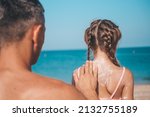 Small photo of A father applies protective cream to his daughter's back at the beach. A man holds sunscreen lotion on a child's body. Cute little girl with sunscreen by the sea. Copy space.
