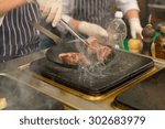 Chef cooking meat in a restaurant in a wok over a commercial griddle or hotplate, close up view of his gloved hands and the meat portions