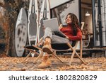 Beautiful woman in cozy outfit works at laptop while sitting on chair near cafe in autumn park.