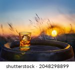 Glass of whisky with ice cubes on old wooden cask. Beautiful sunset at the background.