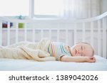 Small photo of Adorable baby girl sleeping in co-sleeper crib attached to parents' bed. Little child having a day nap in cot. Sleep training concept. Infant kid in sunny nursery