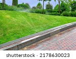 Small photo of automatic lawn watering system in the park with plants, water spray landscape moistening in the backyard with tile pavement and curb, iron grate drainage system, landscaping care theme, nobody.