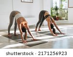 Small photo of Young fit women practice yoga doing asana in a bright yoga studio. Yoga ticher doing asana sun salutation with student in yoga class