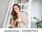 Young woman with brunette long hair in cozy knitted cardigan with cup of tea in hands in bright interior at home