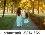 Beautiful young mom walking hand in hand with her young daughter in the park. rear view. Mom and daughter walking down an alley holding hands tightly together. Happy Motherhood