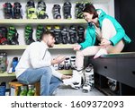 Bearded man helping smiling girlfriend to trying on ski boots in store of sports equipment