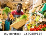 Small photo of Confident african american seller proffering fresh organic carrots and celery in supermarket