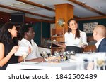 Small photo of positive female busboy serving order to friends in modern restaurante