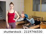 Small photo of Confident active svelte young woman standing in pilates studio, ready for training on reformer machine..