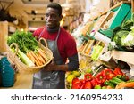 Small photo of Confident african american seller proffering fresh organic carrots and celery in supermarket..