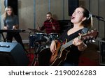 Small photo of Portrait of excited young cheeful girl rock singer with guitar during rehearsal with male drummer and female keyboardist in studio