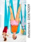 Small photo of Cheerful young girl doing unsupported shoulder stand pose in suspended hammock during group workout of aerial yoga in modern fitness studio