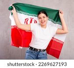 Portrait of young cheerful female in casual wear holding national flag of Iran against gray wall