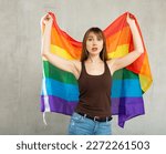 Sorrowful young woman posing in studio against gray background holding colorful rainbow flag of LGBT community