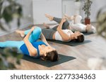 Small photo of Sedulous women doing wind relieving pose of yoga on black mat in light gym room with pot plants